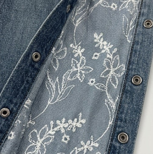 Close-up of denim fabric with floral embroidery and metal buttons.